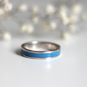 Può includere: Un anello d'argento con un'incastonatura blu brillante. L'anello è posto su uno sfondo bianco, con un elemento floreale sfocato sullo sfondo. La fascia dell'anello è in argento lucido e l'incastonatura blu crea un contrasto sorprendente.