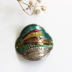 May include: A vintage metal souvenir pin featuring a colorful illustration of Walhalla temple in Regensburg, Germany. The pin has a silver border and the text "WALHALLA REGENSBURG DONAU STAUF". A sprig of dried flowers is in the background.