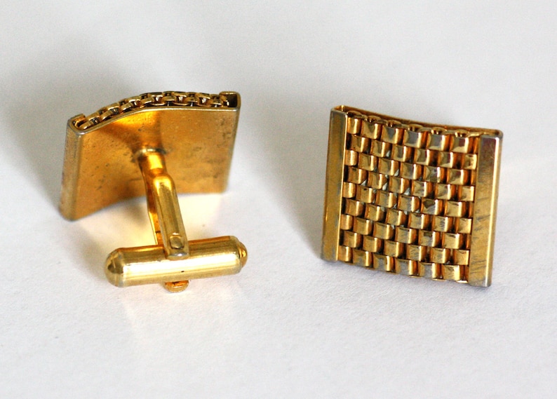 May include: A pair of gold cufflinks with a textured, chain-link design. One cufflink has a traditional back closure, while the other has a more modern, square design.