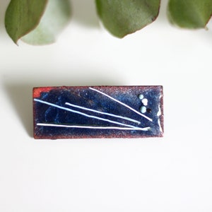 May include: A rectangular enamel pin with a blue background and white lines. The pin has a red border and small white and blue dots.