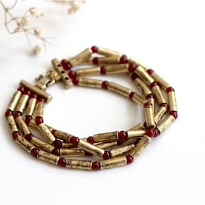 Könnte beinhalten: Eine dreireihige Halskette mit goldfarbenen, zylindrischen Perlen, die mit kleinen, roten, runden Perlen durchsetzt sind. Die Halskette hat einen Verschluss und wird auf einer weißen Oberfläche präsentiert. Die Perlen haben ein leichtes Used-Aussehen.