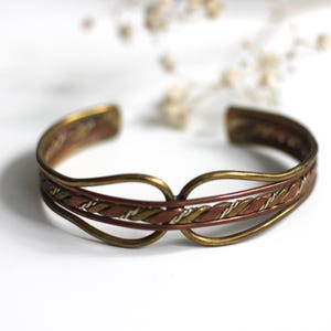 Könnte beinhalten: Ein Vintage-Armreif mit einem Design aus gemischtem Metall. Das Armband hat eine Kombination aus gold-, kupfer- und silberfarbenen Metallen. Das Design umfasst geflochtene und gebogene Elemente. Das Armband wird auf einer weißen Oberfläche präsentiert.