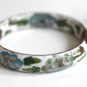 Könnte beinhalten: Ein weißes Emaille-Armreifarmband mit floralem Design. Das Armband hat blaue, grüne und rote Blumen mit grünen Blättern. Der Metallbesatz ist dunkelbraun. Das Armband ist ein Schmuckstück.