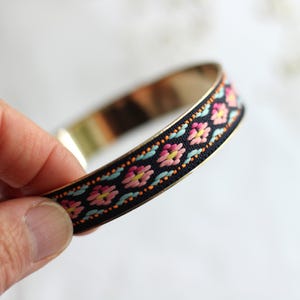 Könnte beinhalten: Ein goldfarbenes Armreif-Armband mit einem schwarzen Band, das ein Blumenmuster in Rosa, Gelb und Türkis aufweist. Das Armband wird in einer Hand gehalten und zeigt das aufwendige Design und die Handwerkskunst des Schmucks.
