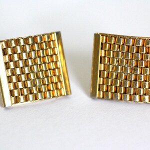 May include: A pair of gold cufflinks with a woven pattern. The cufflinks are square shaped and have a slightly curved design.