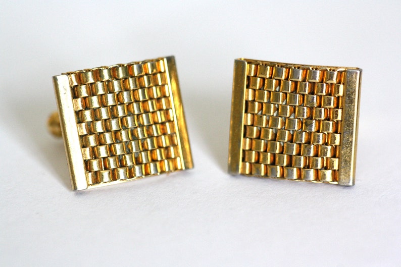 May include: A pair of gold cufflinks with a woven pattern. The cufflinks are square shaped and have a slightly curved design.