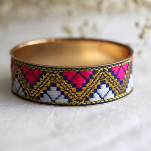 Könnte beinhalten: Ein goldfarbenes Armreif-Armband mit einem farbenfrohen, bestickten Band. Das Band weist ein geometrisches Muster in Rosa, Weiß, Gelb und Blau auf. Das Armband ist ein Schmuckstück.