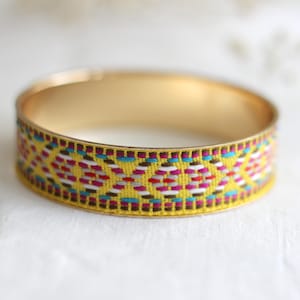 Könnte beinhalten: Ein goldfarbenes Armreif-Armband mit einem gewebten Band mit einem geometrischen Muster in Gelb, Rot, Blau und Weiß. Das Armband hat eine glatte, polierte Oberfläche und ein klassisches Design.