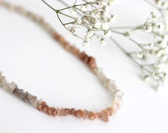 May include: A delicate necklace made of small, irregular, light brown, and gray stones. The stones are strung together on a thin, clear cord. The necklace is lying on a white surface.