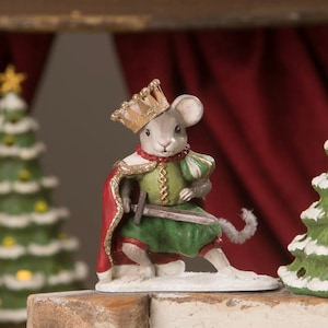 May include: A mouse figurine dressed as a king, wearing a gold crown, red cape, and green tunic, holding a sword. The mouse is posed on a white base, with a Christmas tree in the background. A festive holiday decoration.