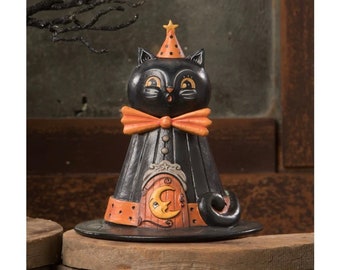 May include: Black cat figurine wearing a witch's hat with a crescent moon door and a star-topped party hat. The cat has a bow tie and is standing on a brown wooden base.