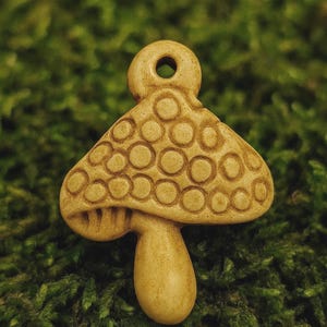 May include: A tan-colored mushroom-shaped pendant with a textured surface and a small hole at the top. The mushroom cap features a pattern of raised circles. The pendant is set against a green, mossy background.