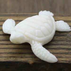 Hand-carved white buffalo bone sea turtle cabochon shown from above, with detailed shell plates and textured flippers outstretched as if swimming, resting on rustic wood as a focal for coastal and beach-inspired jewelry designs.