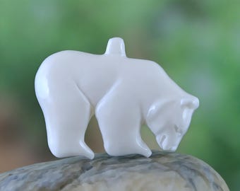 May include: A glossy white polar bear figurine, sculpted in a downward-facing pose. The bear is perched on a textured, gray stone, set against a blurred green background. The figurine has a smooth, reflective surface.