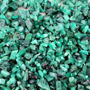 May include: A close-up view of a pile of raw emerald gemstones. The stones are various sizes and shapes, with a vibrant green color and some darker inclusions. The gemstones are scattered, creating a textured surface.