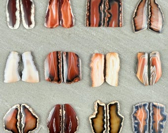 May include: A collection of agate gemstone slices in various shapes and colors. The stones feature bands of brown, orange, white, and gray, with some having a polished edge. The slices are arranged on a gray surface.