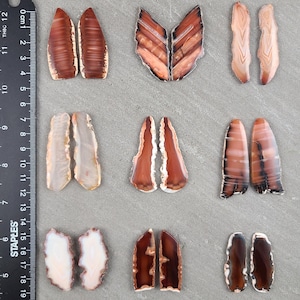 May include: A collection of nine pairs of polished agate slices in various shapes and colors. The stones range from light beige to deep brown, with intricate banding and natural patterns. Each pair is unique, showcasing the beauty of natural stone.