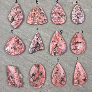 Natural ocean jasper cabochons with pink and coral orbicular patterns and natural druzy pockets arranged on a neutral stone background, each piece uniquely shaped.
