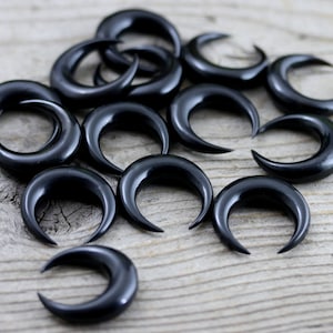 May include: A collection of black crescent moon shaped ear gauges, also known as plugs or tunnels, made of horn. The gauges are arranged in a scattered pattern on a wooden surface.