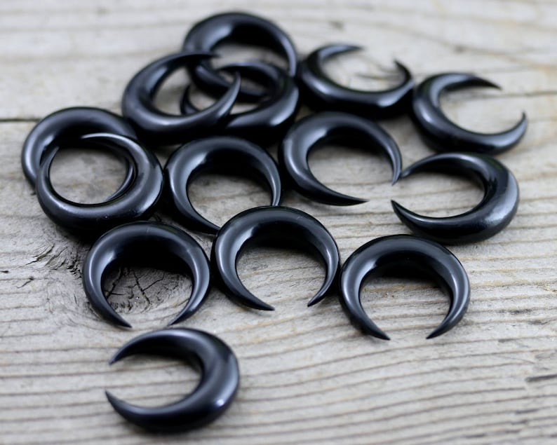 May include: A collection of black crescent moon shaped ear gauges, also known as plugs or tunnels, made of horn. The gauges are arranged in a scattered pattern on a wooden surface.