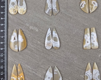 May include: Assortment of polished gemstone pairs in various shapes, including teardrop and elongated forms. The stones feature a mix of creamy white, tan, and brown colors with unique patterns. A ruler in centimeters is visible on the left side.