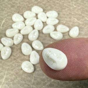 Pode incluir: Uma coleção de pequenas contas brancas em forma oval com um desenho esculpido. Uma conta é segurada por um dedo, mostrando os detalhes intrincados. As contas estão espalhadas sobre uma superfície neutra, sugerindo um tema de artesanato ou joalheria.
