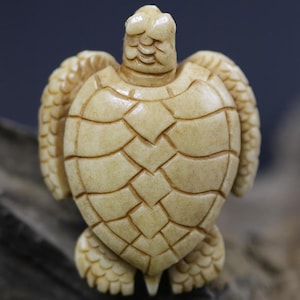 May include: A detailed, cream-colored turtle figurine. The sculpture features a textured shell with geometric patterns, flipper-like limbs, and a detailed head. The turtle is carved from a light-colored material, possibly bone or a similar substance.