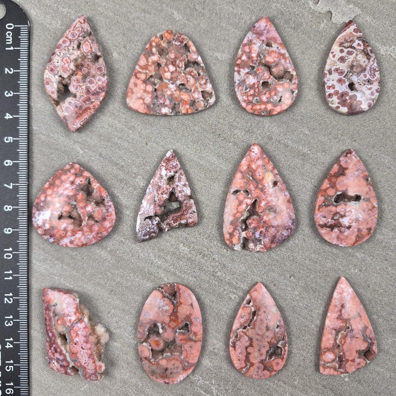 Natural ocean jasper cabochons with pink and coral orbicular patterns and natural druzy pockets arranged on a neutral stone background, each piece uniquely shaped.