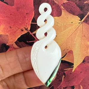 Pode incluir: Pingente branco com design em espiral e base em forma de gota. A base tem um padrão gravado e uma pequena incrustação de concha de abalone. O pingente está sobre um fundo de folhas de outono.