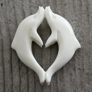 May include: A carved, off-white dolphin pendant. Two dolphins are positioned to appear as if they are kissing, forming a heart shape. The pendant is set against a weathered, gray wooden surface.