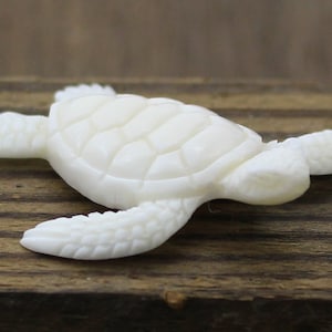 Hand-carved white buffalo bone sea turtle cabochon shown from above, with detailed shell plates and textured flippers outstretched as if swimming, resting on rustic wood as a focal for coastal and beach-inspired jewelry designs.