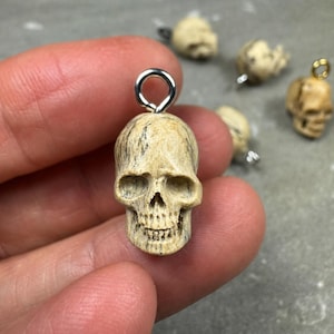 May include: A small, beige, skull-shaped pendant with a textured, wood-grain appearance. The skull features deep eye sockets, a nasal cavity, and detailed teeth. A silver-toned metal jump ring is attached to the top for hanging as a charm.