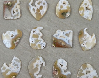 May include: A collection of twelve polished agate gemstones in various shapes, including hearts, teardrops, and irregular forms. The stones feature a mix of beige, white, and brown patterns, with some displaying small cavities and unique veining. The gemstones are arranged on a neutral gray surface.