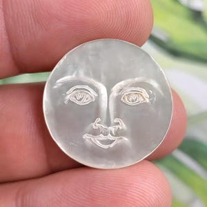 Op de afbeelding: Een witte, ronde, maanvormige hanger met een gegraveerd gezicht. Het gezicht heeft twee ogen, een neus en een mond.