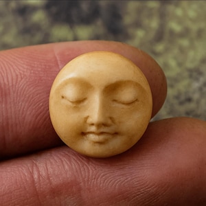 May include: A small, round, beige-colored carving of a serene face with closed eyes. The face has a defined nose and lips, and the overall design suggests a celestial or spiritual theme. The carving is held between two fingers, showcasing its size and detail.