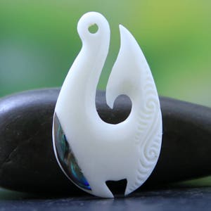 May include: A white bone-like pendant in the shape of a fish hook, with a curved design and a hole at the top. The pendant has a section with iridescent blue and green colors. The hook is resting on a dark gray stone, with a blurred green background.