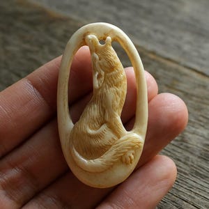 May include: A cream-coloured carved pendant showing a howling wolf inside an oval frame. The wolf has detailed fur and is in a howling pose. The pendant is set against a wooden background. The carving is very detailed.