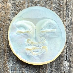 Op de afbeelding: Een ronde, iriserende cabochon met een gesneden maan gezicht. De cabochon heeft een parelmoerglans met tinten blauw, wit en geel. Het gezicht heeft ogen, een neus en een lachende mond. De cabochon staat op een verweerde houten achtergrond.