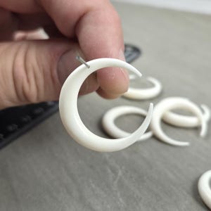 May include: Close-up of several white, crescent-shaped earrings. One earring is held by a hand, with a silver pin inserted. The earrings are smooth and curved. Other earrings are visible in the background, suggesting a jewellery theme.