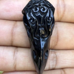 Glossy black, hand-carved stylized raven skull pendant (41mm long) made from buffalo horn, featuring intricate swirling patterns on the cranium.