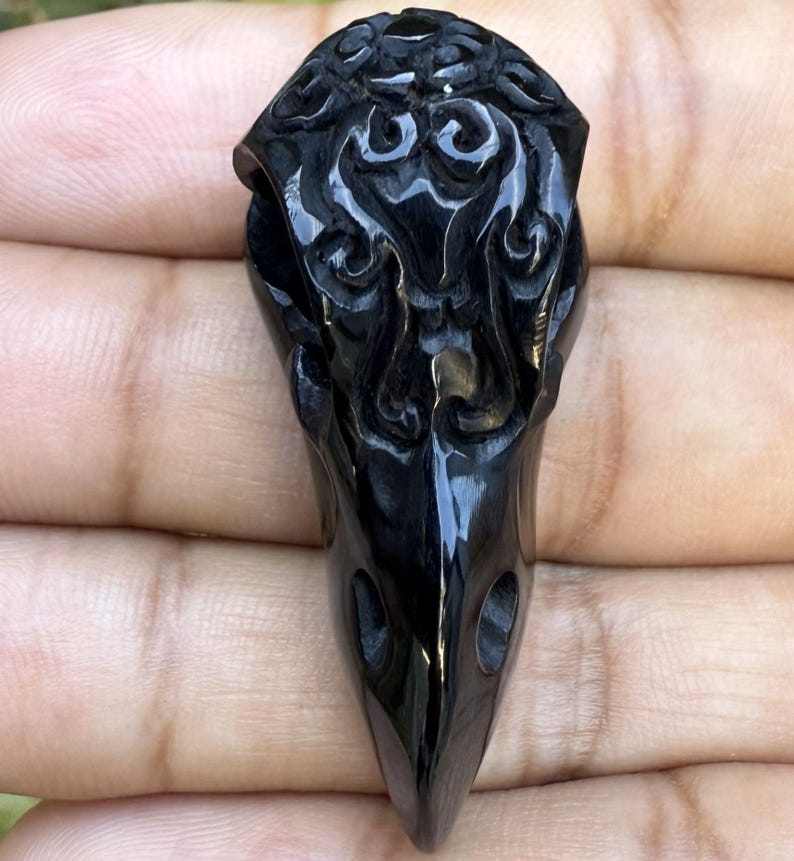 Glossy black, hand-carved stylized raven skull pendant (41mm long) made from buffalo horn, featuring intricate swirling patterns on the cranium.
