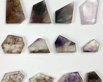 May include: A collection of twelve irregularly shaped, polished gemstones. The stones display a range of colors, including shades of brown, purple, and clear. Each stone has a unique pattern and shape, with some appearing translucent.