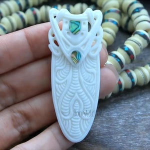 Hand-carved bone pendant featuring intricate tribal designs and two abalone shell inlays.  Elongated shape with a smooth, polished finish. Creamy white bone with iridescent flashes from the abalone. A unique and eye-catching piece of wearable art