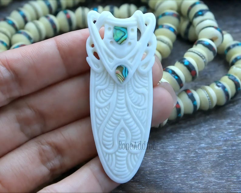 Hand-carved bone pendant featuring intricate tribal designs and two abalone shell inlays.  Elongated shape with a smooth, polished finish. Creamy white bone with iridescent flashes from the abalone. A unique and eye-catching piece of wearable art