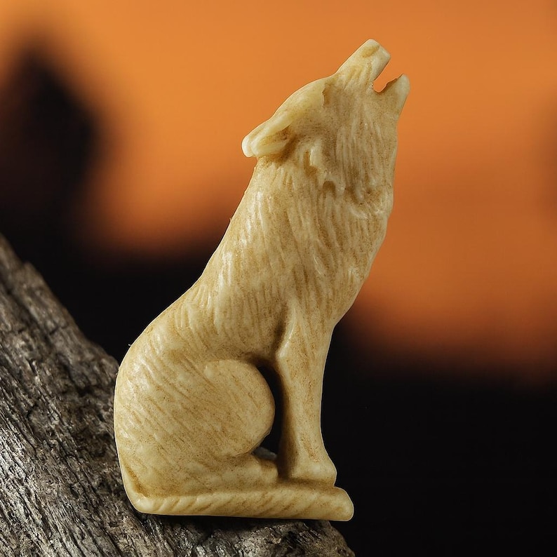 Warm antique-stained bone wolf carving shown in profile on a piece of driftwood, the wolf seated on its haunches with muzzle lifted in a howl, fur texture darkened in the grooves by the stain, flat-backed for setting, wrapping, or stitching, against a soft sunset gradient background, ideal for rustic jewelry, medicine bags, or altar displays.