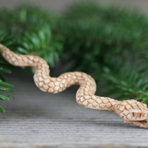 Puede incluir: Una figura de serpiente de madera tallada en color marr&oacute;n claro, con un patr&oacute;n texturizado en forma de escamas. La serpiente est&aacute; colocada en posici&oacute;n curva, con la cabeza ligeramente levantada. Hay agujas de pino verdes en el fondo.