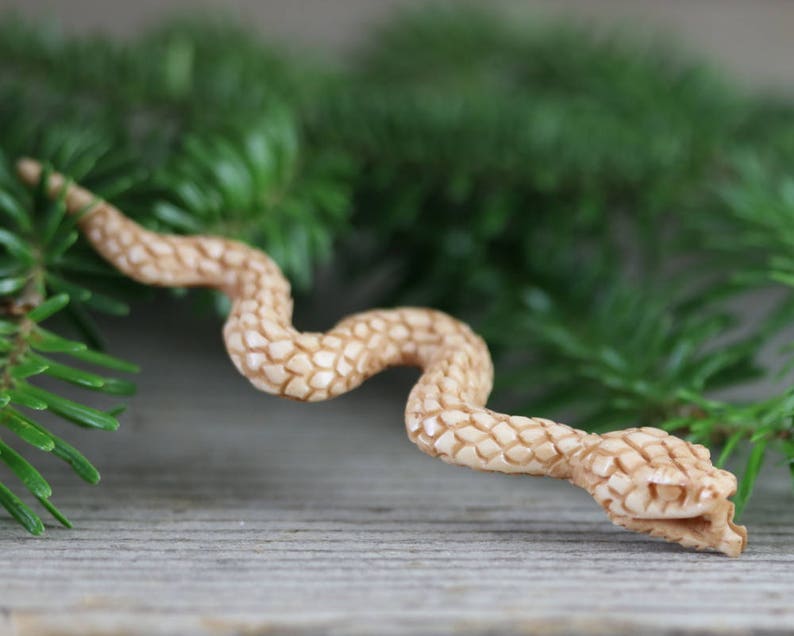 Puede incluir: Una figura de serpiente de madera tallada en color marr&oacute;n claro, con un patr&oacute;n texturizado en forma de escamas. La serpiente est&aacute; colocada en posici&oacute;n curva, con la cabeza ligeramente levantada. Hay agujas de pino verdes en el fondo.