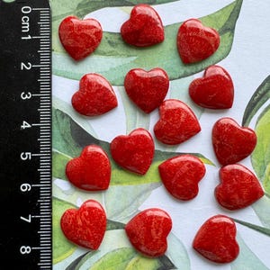 Puede incluir: Una colección de pequeñas cuentas rojas brillantes en forma de corazón. Una regla negra con marcas blancas está en la imagen, mostrando medidas en centímetros. Las cuentas están dispersas sobre una superficie con patrones de hojas verdes y blancas.