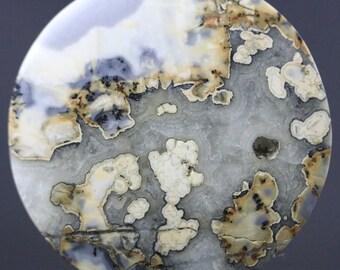 May include: A round, polished stone with a unique pattern. The stone features a mix of colors, including white, gray, tan, and black. The design resembles a landscape with cloud-like formations and intricate details. The stone has a smooth, glossy surface.