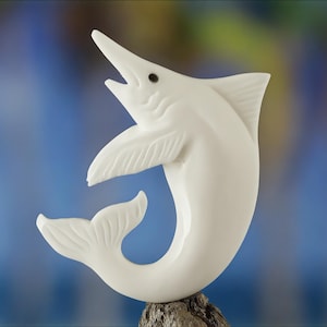 May include: A white sculpture of a marlin fish with an open mouth, fins, and tail. The sculpture is mounted on a piece of wood, with a blurred blue and green background.